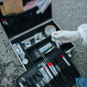 A forensic investigator handles a test tube at a crime scene with an open evidence collection suitcase on the ground.